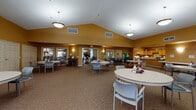 Located at 5863 Scioto Darby Rd in Hilliard, Ohio, this single-floor community facility offers a warm and functional environment designed for group gatherings and shared activities. The centerpiece of the space is a generously sized common room distinguished by soaring vaulted ceilings that lend an open, airy atmosphere. Warm golden-yellow walls create an inviting ambiance, complemented by plush carpeted flooring throughout. The room is illuminated by a combination of pendant light fixtures and recessed ceiling lights, ensuring a well-lit environment. Multiple round tables with modern chairs are arranged throughout the space, accommodating both intimate gatherings and larger group events. Fresh floral centerpieces add a welcoming touch to the dining arrangement. Along one side of the room, a built-in kitchenette with wooden cabinetry and countertop workspace provides convenient food and beverage service. Large windows along the perimeter allow natural light to filter in while offering views of the surrounding area. Additional seating areas and connected rooms are accessible through interior doorways, expanding the functional capacity of the space for a variety of community programs and social activities.