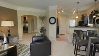 This single-floor residence offers a thoughtfully designed open-concept layout that promotes a sense of spaciousness and connectivity throughout. The living area serves as the heart of the home, featuring plush carpeted flooring, elegant crown molding, and a structural column that adds architectural character without interrupting the flow of the space. A decorative wall clock, vase with tall greenery, and soft neutral wall tones contribute to the polished, welcoming ambiance.

A graceful arched doorway leads to an adjoining room — likely a den or home office — complete with built-in shelving, providing both function and charm. Track lighting overhead ensures the space is well-lit throughout.

Transitioning toward the kitchen, a raised breakfast bar with upholstered bar stools offers a casual dining option. The kitchen features dark countertops that contrast beautifully with the light cabinetry, and a pendant light hangs above, adding a refined finishing touch. A door with a nearby window allows natural light to filter into the kitchen area.

Tile flooring in the kitchen and entry zones contrasts with the soft carpet in the living areas, subtly defining each space. Overall, this residence combines classic architectural details with a warm, neutral palette to create an inviting and functional living environment.