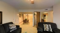 This single-floor residence offers an open and functional layout that seamlessly connects the main living areas. The living room serves as the central gathering space, featuring light beige carpeting, a textured ceiling, and track lighting overhead. Dark upholstered sofas with patterned accent pillows anchor the room, providing a comfortable seating arrangement. An open passage leads directly into the dining area, which is illuminated by a hanging chandelier and accommodates a wooden dining table with matching chairs. Adjacent to the dining space is a laundry area, housing side-by-side washer and dryer units for added convenience. To the right, the kitchen is equipped with wooden cabinetry, a refrigerator, microwave, and additional appliances, offering a practical cooking environment. The continuous carpeting and consistent neutral wall tones throughout create a cohesive and unified feel across all areas. A white paneled entry door is visible near the living room, and HVAC vents are integrated into the ceiling, indicating a centralized climate control system. The overall layout prioritizes accessibility and efficient use of space.