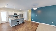 This single-floor, 2-bedroom, 2-bathroom residence offers a thoughtfully designed open-concept layout that seamlessly connects the kitchen, dining, and living areas. The kitchen is a standout feature, equipped with stainless steel appliances — including a refrigerator, microwave, and range — complemented by granite countertops, crisp white cabinetry, and a peninsula with bar seating that makes it ideal for casual dining and entertaining. Pendant lighting above the peninsula and recessed ceiling lights throughout provide a warm, well-lit atmosphere. The living area is distinguished by a bold teal accent wall that introduces a vibrant pop of color against the neutral gray walls and white trim. Durable wood-look flooring runs throughout the main living spaces, while a plush area rug defines the living zone. The layout flows naturally toward the hallway, which leads to the bedrooms and bathrooms, offering a practical and comfortable arrangement. The combination of modern finishes, an open floor plan, and contemporary design details makes this residence a functional and stylish living space well-suited for a variety of lifestyles.