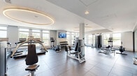 This well-appointed fitness center at 925 S Glebe Rd in Arlington, Virginia, offers a spacious and comprehensively equipped workout facility spread across a single floor. The space is thoughtfully designed with a modern aesthetic, featuring large windows along multiple walls that flood the interior with natural light and provide open views of the surrounding Arlington neighborhood.

The main workout floor is finished with durable large-format dark tiles and is organized into distinct functional zones. A dedicated cardio area houses a row of treadmills, an elliptical trainer, and additional cardio machines, each equipped with individual screens for entertainment. Overhead, striking circular recessed light fixtures add a refined architectural touch to the ceiling.

The strength training zone features a full rack of dumbbells, a power rack with plate-loaded barbells, a flat utility bench, and a cable machine, accommodating a wide range of free weight and resistance exercises. A wall-mounted flat-screen TV keeps users engaged throughout their sessions.

A separate studio area with light hardwood flooring and floor-to-ceiling glass partitions provides a versatile space for stretching, yoga, or group fitness, complete with full-length mirrors. Adjacent to the studio, a clean entry vestibule and restroom facilities with a water fountain ensure added convenience. The overall ambiance is bright, clean, and professional, making this fitness center a standout amenity.