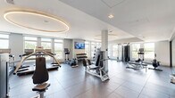 This well-appointed fitness center at 925 S Glebe Rd in Arlington, Virginia, offers a spacious and comprehensively equipped workout facility spread across a single floor. The space is thoughtfully designed with a modern aesthetic, featuring large windows along multiple walls that flood the interior with natural light and provide open views of the surrounding Arlington neighborhood.

The main workout floor is finished with durable large-format dark tiles and is organized into distinct functional zones. A dedicated cardio area houses a row of treadmills, an elliptical trainer, and additional cardio machines, each equipped with individual screens for entertainment. Overhead, striking circular recessed light fixtures add a refined architectural touch to the ceiling.

The strength training zone features a full rack of dumbbells, a power rack with plate-loaded barbells, a flat utility bench, and a cable machine, accommodating a wide range of free weight and resistance exercises. A wall-mounted flat-screen TV keeps users engaged throughout their sessions.

A separate studio area with light hardwood flooring and floor-to-ceiling glass partitions provides a versatile space for stretching, yoga, or group fitness, complete with full-length mirrors. Adjacent to the studio, a clean entry vestibule and restroom facilities with a water fountain ensure added convenience. The overall ambiance is bright, clean, and professional, making this fitness center a standout amenity.