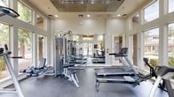 Located at 13015 W Rancho Santa Fe Blvd in Avondale, Arizona, this fitness center offers a bright and inviting environment designed to support a well-rounded exercise routine. The space is defined by its soaring double-height ceilings and floor-to-ceiling windows that wrap around multiple walls, flooding the interior with abundant natural light and providing unobstructed views of the surrounding lush landscaping, palm trees, and outdoor pool area. The open, airy atmosphere is further enhanced by recessed ceiling lighting and a clean, neutral color palette of soft whites and grays throughout.

The exercise room is outfitted with a comprehensive selection of fitness equipment, including a Matrix treadmill, a Concept2 RowERG rowing machine, an elliptical trainer, a multi-station cable strength machine, and an inversion/pull-up tower. A wall-mounted flat-screen television provides entertainment during workouts, while a 24-hour smart vending machine stocked with beverages and snacks adds convenience. The durable dark gray flooring offers a practical, resilient surface suited for a variety of exercises. Double glass doors on one side of the room provide direct access to the outdoor grounds. The overall ambiance is modern, functional, and energizing, making it a well-appointed facility for residents seeking a convenient fitness option.