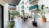 This open-air courtyard serves as a central gathering space, enclosed on multiple sides by a multi-story building with white stucco exteriors and distinctive green-accented balconies. The courtyard features a polished concrete floor that extends the full length of the space, providing a clean and durable surface that complements the surrounding architecture.

A standout focal point is the ornate multi-tiered stone fountain, surrounded by an array of potted tropical plants including palms, ferns, banana trees, and cypress, which lend the space a vibrant, garden-like atmosphere. Additional seating is provided by a bistro-style table and chair set, offering a comfortable spot to relax within the greenery. Benches are also positioned along the perimeter for casual use.

The courtyard is fully open to the sky, allowing abundant natural light to flood the space throughout the day. The surrounding building rises several stories, with each level featuring private balconies fitted with green railings, contributing to a cohesive and visually appealing aesthetic. Ground-floor unit entrances open directly onto the courtyard, creating a seamless connection between interior and exterior spaces. The overall ambiance is tranquil and resort-like, with the dense plantings providing both shade and a sense of lush privacy.