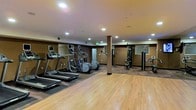 This fitness center is a thoughtfully designed, single-floor facility built to support a comprehensive workout experience. The space features a combination of hardwood flooring and patterned carpet, clearly delineating the cardio and strength training zones. Recessed ceiling lighting illuminates the entire area evenly, creating a bright and energizing atmosphere conducive to exercise.

The cardio section is outfitted with multiple treadmills and elliptical trainers, each equipped with individual display screens for entertainment and performance tracking. A flat-screen television mounted overhead further enhances the cardio experience. The strength training area offers a cable and weight machine setup alongside a dumbbell rack, providing a well-rounded selection of resistance equipment.

Full-wall mirrors line multiple sides of the space, visually expanding the room and allowing users to monitor their form during workouts. Structural columns with warm-toned finishes are integrated into the layout, complementing the neutral color palette of the walls and ceilings. The overall design balances functionality with a clean, professional aesthetic, making this fitness center a practical and inviting facility for daily use.