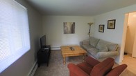 Located at 10 Cedar Blvd in Baldwinsville, New York, this single-floor residence offers a straightforward and functional layout designed for comfortable everyday living. The living area features neutral-toned walls that create a clean, bright backdrop throughout the space. Soft carpeting underfoot adds warmth and comfort to the room. A sturdy pine wood coffee table serves as the centerpiece of the living area, complemented by a plush neutral sofa and a cozy upholstered armchair in a rich rust tone, providing ample seating for relaxing or entertaining. A flat-screen television is positioned on a dark media console, making for a practical and organized entertainment setup. Baseboard heating runs along the walls, ensuring reliable warmth during colder months. Decorative touches, including framed wall art and a floor lamp, contribute to the room's inviting ambiance. The overall atmosphere of the space is calm and unpretentious, with a layout that prioritizes ease of use and livability. Its single-floor design ensures convenient, accessible flow throughout, making it a practical and welcoming home in the Baldwinsville community.
