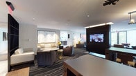 This refined single-floor space is designed with a contemporary aesthetic that balances comfort and functionality throughout its distinct zones. The main lounge area is anchored by plush, neutral-toned sofas and armchairs arranged around low coffee tables, all set atop a bold striped carpet that adds visual depth and movement to the space. Recessed lighting illuminates the area evenly, complementing accent lamps that create a warm, layered ambiance. A wall-mounted flat-screen television is integrated into a sleek dark wood partition, serving as a focal point that divides seating zones while maintaining an open flow.

Curated artwork enhances the interiors throughout, including a striking large-format abstract canvas and a detailed laser-cut city map wall piece that adds a local, artisanal touch to an intimate booth seating nook. Built-in shelving with integrated lighting displays books and decorative objects, lending a residential warmth to what is otherwise a polished, professional environment.

The kitchen area features dark cabinetry contrasted with crisp white countertops, stainless steel appliances, and a large window overlooking lush greenery, flooding the space with natural light. A coffee station and open shelving with a mounted television round out a practical yet stylish service area. Floor-to-ceiling sheer drapery in select seating areas softens the light and connects the interior to outdoor views.