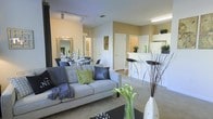 This single-floor residence offers a thoughtfully designed open-concept layout that seamlessly connects the main living spaces. The living room serves as the centerpiece, featuring plush carpeted flooring, track lighting along the ceiling, and ample room for comfortable seating. A stylish sofa accented with decorative pillows anchors the space, complemented by a sleek coffee table adorned with contemporary décor including tall vases and fresh greenery.

Adjacent to the living area, the dining space provides an elegant setting with a mirrored feature that enhances the sense of depth and light throughout the room. The kitchen is positioned toward the rear and boasts a clean, modern design with a raised breakfast bar and bar stool seating, offering both functionality and a casual dining option. Light cabinetry and bright overhead lighting give the kitchen a crisp, welcoming feel.

Warm, neutral-toned walls flow throughout the space, creating a cohesive and inviting atmosphere. Interior doors provide access to additional rooms, suggesting a well-organized floor plan. The overall ambiance is bright and airy, with a contemporary aesthetic that balances comfort and style, making it a versatile and appealing living environment.