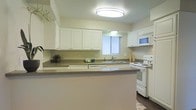 This single-story space showcases a well-organized kitchen as a central feature, designed with practicality and clean aesthetics in mind. The kitchen follows a U-shaped layout enhanced by a peninsula counter, which provides additional workspace and defines the cooking area from adjacent spaces. White raised-panel cabinetry lines the upper walls, offering generous storage while contributing to a bright, cohesive look throughout the space.

Dark laminate countertops provide a subtle contrast against the white cabinetry and appliances, creating a balanced visual appeal. The kitchen is fully equipped with white appliances, including an electric range, an over-the-range microwave, and a dishwasher integrated beneath the countertop. A double-basin sink sits beneath a window dressed with vertical blinds, allowing natural light to filter into the room.

Overhead, a flush-mount ceiling light ensures consistent illumination throughout the kitchen. The flooring transitions from a light-toned material near the kitchen to a darker surface, adding a layer of visual interest. Overall, the space presents a clean, functional environment suited for everyday living.