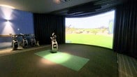 This single-level space is purpose-built as a private indoor golf simulator studio. The room is designed with immersion in mind, centered around a large curved projection screen that wraps across the far wall, displaying realistic golf course visuals with impressive clarity and scale. The curved design of the screen enhances the simulation experience, making it feel as though the player is standing on an actual course.

Dark curtains line the side walls, effectively eliminating light interference and maintaining optimal viewing conditions for the projected imagery. The ceiling is fitted with mounted equipment — likely tracking sensors or projector hardware — that work in tandem with the screen to deliver an accurate simulation experience.

The hitting area features a green artificial turf mat positioned on a carpeted floor, providing a realistic feel underfoot during swings. A full golf bag stocked with clubs is staged at the center of the mat, ready for use, while an additional bag and stand are visible to the side. The overall ambiance is focused and functional, creating a distraction-free environment suited for both casual play and serious practice sessions year-round.