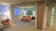 This single-story space showcases a thoughtfully designed bedroom that serves as its central feature. The room is defined by a bold teal accent wall that adds a pop of color against the neutral, warm-toned surrounding walls and ceiling. Soft, wall-to-wall carpeting in a muted beige tone runs throughout, contributing to the room's comfortable and relaxed ambiance.

The bedroom is accessed through a striking slatted wooden sliding partition door, adding a mid-century modern architectural detail that gives the space character and visual interest. A large window fitted with vertical blinds allows natural light to fill the room while offering privacy and light control.

The bedroom is furnished with a full-sized bed dressed in a coral-patterned bedspread and accented with decorative pillows. Flanking the bed are matching nightstands, each topped with a table lamp that provides warm ambient lighting. A trio of framed black-and-white photographs mounted symmetrically on the teal accent wall serves as a focal point, adding a curated, gallery-like touch. The overall ambiance is cozy, organized, and stylistically cohesive.