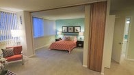 This single-story space showcases a thoughtfully designed bedroom that serves as its central feature. The room is defined by a bold teal accent wall that adds a pop of color against the neutral, warm-toned surrounding walls and ceiling. Soft, wall-to-wall carpeting in a muted beige tone runs throughout, contributing to the room's comfortable and relaxed ambiance.

The bedroom is accessed through a striking slatted wooden sliding partition door, adding a mid-century modern architectural detail that gives the space character and visual interest. A large window fitted with vertical blinds allows natural light to fill the room while offering privacy and light control.

The bedroom is furnished with a full-sized bed dressed in a coral-patterned bedspread and accented with decorative pillows. Flanking the bed are matching nightstands, each topped with a table lamp that provides warm ambient lighting. A trio of framed black-and-white photographs mounted symmetrically on the teal accent wall serves as a focal point, adding a curated, gallery-like touch. The overall ambiance is cozy, organized, and stylistically cohesive.