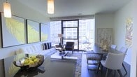 This single-floor urban residence showcases a thoughtfully designed open-plan living and dining space that maximizes both natural light and city views. The centerpiece of the living area is a dramatic floor-to-ceiling window wall framed in dark metal, flooding the interior with daylight and providing direct sightlines to the surrounding metropolitan skyline. A sliding glass door connects the interior to a private outdoor terrace, ideal for al fresco relaxation.

The living area is anchored by a plush white sofa accented with navy throw pillows, arranged around a sleek, low-profile coffee table atop a richly patterned area rug. A modern armchair complements the seating arrangement, while large-scale artwork adorns the walls, adding color and visual depth to the space. Pendant and table lighting provide warm, layered illumination throughout.

Seamlessly connected to the living space, the dining area features a dark-toned table with contemporary upholstered seating. Decorative elements, including sculptural accents and botanical artwork, reinforce the sophisticated, cosmopolitan aesthetic that defines this well-appointed urban residence.