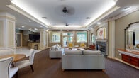 This single-story community facility is thoughtfully designed to provide a variety of functional and comfortable gathering spaces. The main lounge serves as the centerpiece, featuring tray ceilings with ambient cove lighting, ceiling fans, and rich patterned carpeting throughout. A marble-surround fireplace anchors one wall, flanked by decorative arched niches with ornamental mirrors and shelving. Large glass doors and windows along the exterior wall flood the space with natural light and provide views of the outdoor greenery, while a seating arrangement of neutral gray sofas and accent chairs in warm yellow tones creates an inviting atmosphere.

Adjacent to the main lounge is a dedicated media room, highlighted by a stone-framed entertainment center with built-in cabinetry, a flat-screen television, and a surrounding bank of backlit glass panels. Built-in shelving and a glass-top coffee table complete the room's polished, functional aesthetic.

A reception area features a wraparound granite countertop with dark espresso cabinetry and a bay window, providing a welcoming point of entry. A separate meeting or game room offers multiple round tables surrounded by upholstered chairs, with natural light filtering through wood-slatted blinds. Together, these spaces create a cohesive, elegantly finished environment well-suited for community gatherings and social activities.