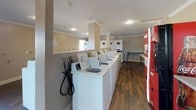 Located at 1539 Tavern Road in Alpine, California, this single-story community laundry facility serves as a convenient shared amenity set within a lush, tree-lined residential environment. The exterior presents a stucco finish complemented by mature trees, manicured shrubs, and well-kept walkways that create an inviting approach to the building.

Inside, the laundry facility is thoughtfully laid out to accommodate high-volume use. A generous row of top-load washing machines lines one wall, while stacked dryer units occupy the opposite side, providing ample capacity for residents. The interior features durable hardwood-style flooring, neutral-toned walls, and crown molding details that lend a clean, polished aesthetic. Recessed lighting keeps the space bright and functional throughout the day. A central support column divides the room subtly without disrupting workflow. A vending machine is conveniently positioned near the entrance, and a utility table offers additional surface space for folding or sorting laundry.

The surrounding grounds feature abundant mature trees, layered landscaping with a variety of shrubs and plants, and paved walkways with wooden railings, all contributing to a tranquil, well-maintained outdoor setting that enhances the overall appeal of the facility.