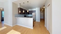 This single-floor residence features a thoughtfully designed open layout that connects the kitchen and living areas seamlessly. The living room is bright and airy, with light wood flooring, neutral-toned walls, and large windows that allow generous natural light to filter in, casting warm patterns across the floor.

The kitchen is a standout space, showcasing rich dark-wood cabinetry that extends to the ceiling for ample storage, complemented by granite countertops with subtle blue-grey tones. A built-in tile backsplash adds texture, while stainless steel appliances—including a refrigerator, range, microwave, and dishwasher—provide a polished, modern aesthetic. A peninsula with pendant lighting overhead offers additional prep space and casual seating.

The bedroom is a calm, carpeted retreat featuring a closet and a sizable window with views of surrounding greenery, filling the room with natural light.

The bathroom is well-appointed with a long granite-topped vanity, dark wood cabinetry with brushed nickel hardware, dual sinks, and a large mirror. A stylish multi-light fixture provides ample illumination, while soft carpeted flooring adds warmth. The overall ambiance of the space is clean, contemporary, and move-in ready.