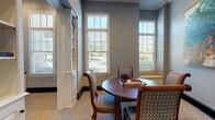 This thoughtfully designed single-floor space offers a refined blend of function and comfort, finished in a cohesive cool-gray palette throughout. The central lounge and meeting area anchors the space with a rich mahogany round table surrounded by elegantly upholstered chairs featuring a classic dot pattern. A bold Greek key-patterned carpet runs throughout, adding visual interest underfoot while tying the rooms together seamlessly. Multiple double-hung windows dressed with white shutters flood the lounge with abundant natural light and offer pleasant views of lush greenery outside. Built-in white bookshelves with cabinetry provide both storage and architectural character, while a white credenza topped with a coffee station and beverage accessories adds a welcoming hospitality touch. Colorful framed artwork adorns the walls, providing vibrant focal points against the muted gray walls. A dedicated business center features a long built-in desk with upper cabinetry, accommodating multiple workstations equipped with desktop computers and ergonomic task chairs, alongside a window that brings in natural light. French glass-paned doors open to a fitness room visible in the background, housing cardio equipment. The space is further enhanced by clean white trim work, crown molding, and dark baseboards that frame each room with a polished, traditional finish.