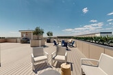 Located at 9191 Nelson Rd in Longmont, Colorado, this rooftop deck offers an impressive open-air retreat designed for relaxation and social gatherings. The deck is surfaced with light-toned composite wood planking that extends across a generously sized, rectangular layout bordered by low perimeter walls lined with lush greenery and plantings.

Throughout the space, oversized raised planters serve as natural focal points, each housing mature trees that provide shade and a touch of nature against the open Colorado sky. The deck is organized into several distinct seating zones, including clusters of white lounge chairs, Adirondack-style chairs in bold navy blue, built-in bench seating, and dining table sets — offering a variety of settings for both casual lounging and group dining.

A dedicated grilling and outdoor kitchen area is equipped with gas grills and prep stations, making the space well-suited for outdoor entertaining. Waste and recycling stations are thoughtfully integrated into the layout. The perimeter greenery hedge adds privacy while framing breathtaking panoramic views of the Rocky Mountain foothills and the expansive Colorado landscape beyond. Under wide-open skies, this rooftop deck delivers a bright, airy ambiance that perfectly captures the spirit of Colorado outdoor living.