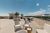 Located at 9191 Nelson Rd in Longmont, Colorado, this rooftop deck offers an impressive open-air retreat designed for relaxation and social gatherings. The deck is surfaced with light-toned composite wood planking that extends across a generously sized, rectangular layout bordered by low perimeter walls lined with lush greenery and plantings.

Throughout the space, oversized raised planters serve as natural focal points, each housing mature trees that provide shade and a touch of nature against the open Colorado sky. The deck is organized into several distinct seating zones, including clusters of white lounge chairs, Adirondack-style chairs in bold navy blue, built-in bench seating, and dining table sets — offering a variety of settings for both casual lounging and group dining.

A dedicated grilling and outdoor kitchen area is equipped with gas grills and prep stations, making the space well-suited for outdoor entertaining. Waste and recycling stations are thoughtfully integrated into the layout. The perimeter greenery hedge adds privacy while framing breathtaking panoramic views of the Rocky Mountain foothills and the expansive Colorado landscape beyond. Under wide-open skies, this rooftop deck delivers a bright, airy ambiance that perfectly captures the spirit of Colorado outdoor living.