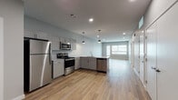 This thoughtfully designed single-floor unit offers a bright and contemporary living experience anchored by an open-concept kitchen and living area. The kitchen is outfitted with a full suite of stainless steel appliances, including a refrigerator, gas range, built-in microwave, and dishwasher. Gray shaker-style cabinetry pairs with light stone countertops, while an L-shaped peninsula provides additional prep space and casual seating. Stylish pendant lights hang above, adding a modern touch to the space. Light-toned wood-style flooring flows seamlessly throughout the entire unit, enhancing the sense of continuity and openness. The living area benefits from natural light streaming through large windows, creating an airy and welcoming ambiance. The bedrooms are generously proportioned, each featuring neutral-toned walls, ceiling fans, and windows fitted with blinds. Closet storage is included in the bedrooms. The bathroom is finished with a classic white tiled tub-shower surround with a built-in niche, complemented by a gray vanity with stone countertop and chrome fixtures. Recessed lighting throughout the unit contributes to a clean, modern aesthetic across every room.