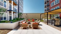 This expansive outdoor courtyard is set within the interior of a modern multi-story residential complex, characterized by a striking façade that blends bold red and neutral gray siding with large windows and private balconies. The courtyard is thoughtfully designed to offer a range of outdoor living experiences across a single level.

At its heart, a generous paved terrace of large-format light stone tiles provides a clean, open gathering area. Multiple seating arrangements feature teak-framed chairs with vibrant red cushions and low-profile sofas with neutral upholstery, all oriented around a sleek fire table — ideal for relaxed social gatherings. A patch of well-maintained lawn offers a softer counterpoint to the hardscape surfaces nearby.

A pergola-covered outdoor kitchen and grilling station anchors one end of the courtyard, equipped with built-in grills and bar-height dining seating beneath a canopy that provides welcome shade. Hammocks add a leisure-focused element to another corner of the space.

A naturalistic garden path winds through a section of the courtyard, featuring stepping stones set among river rock and ornamental plantings, lending a serene, park-like quality. Raised planting beds line the perimeter, filled with grasses, shrubs, and young trees that soften the surrounding architecture. The open sky above floods the entire space with natural light throughout the day.