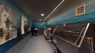 This single-floor pet grooming facility is thoughtfully designed to provide a functional yet visually engaging environment for pet care. The room is laid out in a long, narrow configuration, maximizing the use of space along one primary wall. Multiple stainless steel wash tubs are positioned in a row, each equipped with handheld showerheads, grooming arm attachments, and integrated fixtures, offering dedicated stations for bathing and grooming pets of various sizes.

The walls are adorned with vibrant ocean-themed murals depicting rippling water, creating a cohesive aquatic ambiance throughout the space. Large-format photographic prints of dogs—including a Saint Bernard, a white German Shepherd, and others—are displayed prominently, adding character and warmth to the room. The lower portion of the walls is finished with dark blue ceramic tile, which transitions seamlessly into the matching tile flooring, ensuring durability and ease of cleaning.

Overhead, a dark ceiling features sleek linear LED strip lighting running the length of the room, complemented by recessed downlights that provide even, practical illumination. A mobile utility cart stocked with pet care products and a professional dryer are also present. A glazed entry door provides access, completing this well-equipped, purpose-built grooming space.