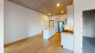 Located at 333 E Hennepin Ave in Minneapolis, this modern unit showcases a striking industrial-chic aesthetic defined by exposed concrete ceilings with visible ductwork that contrast beautifully against clean white walls and warm wood-tone flooring throughout. The open-concept kitchen and living area serves as the heart of the space, featuring a central island with a sleek white quartz countertop, flat-front cabinetry in both light wood and white finishes, a full suite of stainless steel appliances including a gas range, refrigerator, dishwasher, and microwave, and a tile backsplash. Recessed lighting illuminates the kitchen, while the living area benefits from abundant natural light streaming through large windows and a glass sliding door that opens to a private balcony with expansive views of the Minneapolis skyline. The bedroom is generously proportioned with the same wood-look flooring, exposed ceiling details, and direct balcony access, filling the room with natural light and city views. The bathroom is finished in a clean, minimalist style with a wide vanity topped with an integrated white sink, light wood cabinetry, a backlit mirror, a combined tub and shower, and durable wood-tone flooring. The overall ambiance of this space is contemporary and airy, blending urban industrial character with modern comfort.