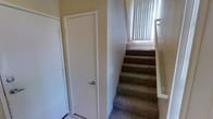 This space showcases a clean, functional interior design with a warm and neutral color palette throughout. The carpeted staircase is a central feature, fitted with a sturdy handrail and topped by a window dressed with vertical blinds that allows natural light to filter in, brightening the stairwell and creating a welcoming atmosphere. The walls carry a soft, warm beige tone complemented by white trim and door frames, contributing to a cohesive and tidy aesthetic. The lower landing area features light-colored tile flooring, providing a practical transition point between levels. A white interior door with a brushed silver lever handle is conveniently located near the base of the stairs, offering easy access to an adjacent room or utility space. The textured wall surfaces add subtle visual depth to the interior. Overall, the space presents a straightforward, well-kept layout that balances practicality with a comfortable residential feel, making efficient use of the available area while maintaining a bright and inviting atmosphere through thoughtful use of natural light and neutral finishes.