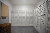 This dedicated mail room is designed to efficiently serve a large multi-unit building, offering a centralized and well-maintained space for resident mail collection. The room is lined on multiple walls with an extensive bank of individual, lockable metal mailboxes in a neutral gray finish, accommodating a significant number of occupants. The mailbox units are arranged in tall, multi-row configurations and include larger parcel compartments at the lower sections, ensuring versatility for various mail and package sizes. The flooring features a patterned, low-pile carpet in muted blue and gray tones, adding a subtle design element to the otherwise functional space. The walls are finished in a clean, light gray tone that contributes to a bright and tidy atmosphere, complemented by ceiling-mounted lighting that evenly illuminates the room. A wicker basket is positioned near one of the mailbox units, providing a practical touch for residents. A cork bulletin board is mounted near the exit, offering a space for community notices. A fire extinguisher is visibly mounted in the hallway just beyond the room, reflecting attention to safety. The layout allows for easy circulation, with a connecting hallway providing smooth access to other areas of the building.