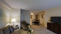 This single-story residence features an open-concept layout that seamlessly connects the living and dining areas, creating a cohesive and functional living space. The living area is appointed with light-toned carpet flooring and warm beige walls, contributing to a comfortable and inviting ambiance. A decorative floor lamp provides soft accent lighting, complementing the natural and overhead light sources throughout the space.

The dining area is positioned adjacent to the living space and is outfitted with a ceiling fan, offering both lighting and air circulation. A dark-finished dining table and matching chairs anchor the space, offering a defined area for meals. A pass-through opening in the wall connects the dining area to what appears to be the kitchen, providing convenient access and an added sense of openness.

A louvered closet door is visible along one wall, suggesting practical built-in storage solutions. The neutral color palette of warm beige and taupe tones is consistent throughout, lending the space a unified and understated aesthetic. Overall, the layout prioritizes ease of flow between living and dining functions, making it a practical and welcoming environment.