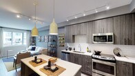 This thoughtfully designed single-floor space showcases a contemporary open-concept layout that seamlessly integrates the kitchen and living area into one cohesive environment. The kitchen is a standout feature, outfitted with warm grey wood-finish cabinetry, a crisp white subway tile backsplash, and a suite of stainless steel appliances including a range, built-in microwave, and dishwasher. A generous white countertop island anchors the space, providing both prep space and casual seating, accented with woven placemats and decorative accessories that add a warm, curated touch. Pendant lighting in a soft yellow-cream finish hangs above the island, complementing the track lighting system that runs along the ceiling to provide ample, well-distributed illumination throughout. The living area, visible beyond the kitchen, is furnished with a comfortable sofa and accented by an open industrial-style shelving unit, creating a relaxed yet modern atmosphere. Soft blue-grey wall tones lend a calm, airy quality to the interior, while the open-plan design maximizes the sense of space and encourages a fluid, connected lifestyle. The overall aesthetic balances modern functionality with warm, inviting design elements.