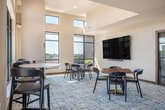 Located at 517 W Washington Ave in the heart of Madison, Wisconsin, this thoughtfully designed communal space combines a kitchen, dining area, and living room in a seamless open-plan layout under impressively high ceilings that amplify the sense of openness throughout.

The kitchen serves as a vibrant focal point, anchored by a striking teal island with a white marble countertop and seating for multiple guests. Overhead, hammered brass pendant lights add warmth and character. The back wall features navy cabinetry with brass hardware, arched tile-backed niches with open shelving, and a luminous neon lotus art piece that lends the space a distinctive, contemporary personality.

The dining area transitions naturally from the kitchen, offering both standard and bar-height seating options arranged to take full advantage of the floor-to-ceiling black-framed windows. These expansive windows flood the space with natural light and frame panoramic views of the Madison skyline.

The living area is furnished with modern lounge seating, a sculptural round coffee table, and a richly patterned area rug over light hardwood flooring, creating a comfortable and inviting atmosphere. A wall-mounted flat-screen TV and glass doors opening to an outdoor rooftop terrace complete the space. The rooftop terrace itself features sectional seating, planters with greenery, and unobstructed views of the surrounding cityscape.