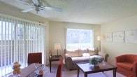 This single-floor space offers a warm and inviting atmosphere, thoughtfully designed for comfortable everyday living. The living area serves as the heart of the home, featuring a ceiling fan overhead and a textured ceiling that adds subtle character. Large windows dressed with vertical blinds flood the room with abundant natural light, creating an airy and open feel throughout. A sliding glass door provides seamless access to the outdoors, further enhancing the sense of openness and connectivity to the exterior environment.

The living room is well-appointed, with ample space for a plush sofa set and a coffee table, making it an ideal area for relaxing or entertaining. Neutral-toned walls and light carpeting contribute to the space's warm, understated aesthetic. The dining area sits adjacent to the living room, accommodating a dining table and chairs, and benefits from the same natural light that permeates the space. Together, the living and dining areas form a cohesive, open-plan layout that feels both functional and welcoming. Overall, this space strikes a balance between practicality and comfort, making it a pleasant environment for day-to-day living.