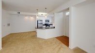 This single-floor residence is designed with a functional, open layout that connects the kitchen and living area seamlessly. The kitchen is a standout feature, outfitted with white cabinetry, dark granite countertops, and a full suite of stainless steel appliances including a refrigerator, gas range, and built-in microwave. A peninsula with matching granite provides additional counter space and casual seating, while two pendant lights hang above, adding focused illumination. Hardwood flooring runs through the kitchen, transitioning to plush carpeting in the living area, which is bright and open with track lighting along one wall.

The bedroom is a comfortable, carpeted space with a ceiling fan and a window with blinds that brings in natural light and a view of greenery outside. A hallway connects the bedroom to the bathroom, which features white tile flooring, a tub-shower combination, a wide vanity with dark granite countertops, white cabinetry, and dual mirrors above. A utility panel is accessible from the hallway. The entry door includes a transom window above, adding natural light near the front of the unit. Throughout, the space maintains a clean, neutral palette with white walls and bright trim.
