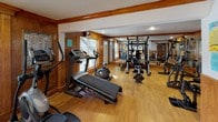 This single-floor fitness space is thoughtfully designed to accommodate a full range of workout activities. The room features warm wood-paneled wainscoting along the walls, complemented by light hardwood flooring that creates a cohesive, inviting aesthetic. Structural beams with wood trim detail the ceiling, adding architectural character throughout the space.

The fitness area is generously sized, providing ample room for a diverse array of equipment. A Gold's Gym multi-station cable machine serves as a central strength-training hub, accompanied by a dedicated weight bench, free weights, and various resistance machines. Cardio equipment includes a treadmill, elliptical trainer, and rowing machine, offering versatile options for aerobic exercise.

Large windows along one wall allow natural light to filter in, brightening the workout area, while wall-mounted mirrors expand the sense of space and allow for form monitoring during exercise. A wall-mounted television is also present for entertainment during workouts. A French door leads to an adjoining room, enhancing the overall flow of the space. The textured ceiling and recessed lighting complete the functional yet comfortable atmosphere of this well-appointed fitness facility.