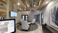 This thoughtfully designed single-floor space makes an immediate impression with its dramatic vaulted ceiling, where exposed wood beam trusses and white-painted structural elements create an open, airy atmosphere. Track lighting and recessed fixtures illuminate the space effectively, highlighting the warm wood tones and modern aesthetic.

The central lounge area serves as the focal point, featuring a curated seating arrangement of contemporary upholstered chairs positioned around a round accent table atop a circular patterned area rug with bold, flowing lines. Wood-look tile flooring in a neutral tone runs throughout, adding warmth and visual continuity.

The walls are finished in a soft gray palette that complements the natural wood ceiling accents. A large flat-screen display is mounted on one wall, while a dark credenza provides functional storage nearby. Double glass-paned doors at the rear offer a view to the exterior, allowing natural light to filter into the space.

Additional rooms are accessible through doorways along the side wall, suggesting supplementary functional areas beyond the main lounge. The overall ambiance is polished and welcoming, blending rustic architectural elements with clean, modern furnishings to create a space that is both professional and comfortable.