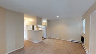 This single-floor unit offers a clean, functional layout with an open-concept design that connects the kitchen and living area seamlessly. The kitchen is equipped with white appliances, including a full-size refrigerator, and features warm wood-toned cabinetry. A peninsula-style breakfast bar serves as a natural divider between the kitchen and the living space, providing a practical surface for casual dining or food preparation. The living area is finished with wall-to-wall carpeting in a neutral beige tone, contributing to a warm and understated ambiance. Walls are painted in a soft, light neutral color, creating a bright and airy atmosphere throughout. Recessed lighting in the kitchen adds focused illumination, while a baseboard heating unit along the living room wall ensures efficient climate control. The textured ceiling adds a subtle architectural detail consistent throughout the space. Overall, the unit presents a straightforward and practical interior design with a cohesive neutral palette, making it a versatile space well-suited for comfortable, everyday living.