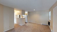 This single-floor unit offers a clean, functional layout with an open-concept design that connects the kitchen and living area seamlessly. The kitchen is equipped with white appliances, including a full-size refrigerator, and features warm wood-toned cabinetry. A peninsula-style breakfast bar serves as a natural divider between the kitchen and the living space, providing a practical surface for casual dining or food preparation. The living area is finished with wall-to-wall carpeting in a neutral beige tone, contributing to a warm and understated ambiance. Walls are painted in a soft, light neutral color, creating a bright and airy atmosphere throughout. Recessed lighting in the kitchen adds focused illumination, while a baseboard heating unit along the living room wall ensures efficient climate control. The textured ceiling adds a subtle architectural detail consistent throughout the space. Overall, the unit presents a straightforward and practical interior design with a cohesive neutral palette, making it a versatile space well-suited for comfortable, everyday living.