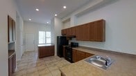 This single-floor space makes a strong impression with its open-concept kitchen, designed to be both functional and visually spacious. The kitchen is outfitted with warm, rich medium-toned wood cabinetry that extends from counter height all the way up to the tall ceiling, maximizing storage capacity and drawing the eye upward to emphasize the generous vertical space. A full suite of black appliances, including a refrigerator and electric range with overhead ventilation, anchors the cooking area, complemented by light-toned laminate countertops that run along the perimeter. A central kitchen island provides additional prep and storage space, while a stainless steel sink adds a functional finishing touch. Recessed lighting is installed throughout the ceiling, providing bright, even illumination across the entire space. Large tile flooring in a neutral beige tone runs throughout, offering durability and a clean, cohesive look. A window near the entry door allows natural light to filter in, enhancing the airy feel of the room. The tall ceilings and open layout combine to create a sense of openness and ease of movement throughout this well-appointed space.