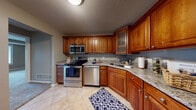 This single-floor residence offers a thoughtfully designed layout that balances functionality and comfort throughout its well-maintained spaces. The kitchen serves as a standout feature, showcasing rich, warm-toned wood cabinetry with raised-panel doors and glass-front accent cabinets. Granite countertops in a speckled neutral palette provide ample workspace, while a full suite of stainless steel appliances — including an electric range, built-in microwave, dishwasher, and double-basin sink — delivers a complete cooking experience. Light tile flooring grounds the kitchen and transitions naturally into the adjoining hallway and entry areas. A half-wall pass-through with a granite ledge connects the kitchen to the main living area, fostering an open and communicative flow between spaces. The living area is generously proportioned, featuring soft wall-to-wall carpeting, neutral taupe walls, and textured ceilings. Natural light filters in through windows visible along the perimeter. The bathroom includes a full soaking tub and shower combination set against beige tile surround. A dedicated in-unit laundry closet houses a stacked washer and dryer, adding everyday convenience. Closet storage and carpeted bedroom spaces complete this practical, move-in-ready home.