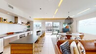 This stylish single-floor space showcases a contemporary open-plan design that seamlessly integrates the kitchen, dining, and living areas into one cohesive, light-filled environment. Large-format tile flooring runs throughout, lending a clean and expansive feel to the interior.

The kitchen is thoughtfully appointed with white cabinetry, a subway tile backsplash, open wood shelving, a gas range, and a generous island clad in warm wood with a dark countertop and gold-toned bar seating. A striking yellow wood-slat ceiling detail above the kitchen and dining transition zone adds a bold architectural accent.

The dining area connects fluidly to a spacious living room, where oversized windows frame sweeping views of the surrounding rooftops and distant mountain ranges. The living room is bathed in natural light, creating a warm and inviting atmosphere. A hallway with a warm wood ceiling and full-length windows further enhances the sense of brightness throughout.

Sliding glass doors open directly onto a generous outdoor terrace, complete with outdoor seating and a gas grill, offering a seamless indoor-outdoor living experience. The overall ambiance of the space is modern and airy, characterized by a refined material palette of warm wood tones, crisp white walls, and large glazed openings that maximize natural light and views.