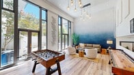 Located at 2425 Prospect St in Berkeley, CA, this single-floor communal living space offers a thoughtfully designed environment that balances relaxation, socializing, and productivity. The room is defined by soaring double-height ceilings that lend an open, airy quality throughout, complemented by wide-plank hardwood floors that run the length of the space.

At the heart of the room, floor-to-ceiling glass doors and tall windows flood the interior with abundant natural light and provide direct views to an outdoor courtyard area. A striking abstract mural in deep blue tones anchors one wall, while a bold black-and-white graphic artwork commands attention on another, giving the space a gallery-like character.

The lounge area is furnished with a tan leather sofa, curved accent chairs, and a low coffee table, creating a comfortable gathering spot. A foosball table adds a recreational touch to the open central zone, emphasizing the space's social character. Along one wall, a row of built-in desk nooks with individual lighting provides dedicated work or study stations, making the space equally suited for focused tasks.

A secondary seating alcove with additional lounge furniture and a credenza offers a quieter retreat within the larger room. Lush potted tropical plants and curated decorative wall installations complete the contemporary aesthetic, resulting in a vibrant, well-appointed communal space.