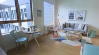 This thoughtfully designed single-floor space exudes a warm, Scandinavian-inspired aesthetic that balances simplicity with comfort. The open-plan layout seamlessly connects a casual dining area with a relaxed living zone, creating a cohesive and inviting atmosphere throughout.

The dining area features a compact round wooden table paired with mint-green metal chairs, lending a playful pop of color against the clean white walls. Large windows on multiple sides flood the space with abundant natural light, enhancing the airy, open feel and creating a bright, welcoming environment throughout the day.

The living area is furnished with a plush grey sofa accented with patterned cushions, a woven rattan coffee table, and a sculptural accent chair in natural wood tones. A geometric multicolored area rug anchors the seating arrangement, adding a lively visual element to the light hardwood flooring that runs throughout the space.

Thoughtful lighting is provided by a minimalist tripod floor lamp, complementing the natural light from the oversized windows. Framed artwork on the walls adds personality and depth without overwhelming the clean, uncluttered design. The overall ambiance is calm, modern, and casually sophisticated — ideal for both everyday living and relaxed entertaining.