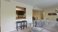 This single-floor residence showcases a thoughtfully designed open-concept layout that promotes a seamless flow between living, dining, and kitchen areas. The living area features neutral-toned carpet flooring and is furnished with a plush gray sofa, creating a comfortable and inviting atmosphere. A standout architectural detail is the pass-through kitchen window, framed cleanly into the wall and accented with a granite countertop ledge. This practical feature connects the kitchen to the living space while also serving as a casual bar area, complemented by two black bar stools. The kitchen itself, visible through the pass-through, is equipped with dark cabinetry and modern appliances. Beyond the living area, a dining space is visible, featuring a dark wood table and chairs, maintaining the cohesive, neutral color palette found throughout the home. A hallway leading from the main living area suggests additional rooms within the residence. The walls are painted in a warm, neutral tone, contributing to the bright and airy feel of the space. Overall, the layout is efficient and well-connected, making it an ideal space for both everyday living and entertaining.