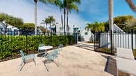 Located at 10471 SW 216th St in Cutler Bay, Florida, this community outdoor amenity space offers a well-appointed pool and recreation area designed for relaxed, resort-style living in South Florida. The centerpiece is a generously sized swimming pool with crystal-clear turquoise water, complemented by a smooth, wide pool deck that provides ample room for sunbathing and socializing. White lounge chairs are positioned along the deck for leisure, while aqua-toned bistro tables and chairs are arranged throughout for casual outdoor dining and conversation. A covered pavilion with sturdy concrete columns offers a shaded retreat from the Florida sun, furnished with seating that makes it ideal for gatherings. The area also features a dedicated lounge section with additional seating surrounded by mature palm trees and tropical greenery, creating a private, serene atmosphere. The space is enclosed by a sleek black iron fence, ensuring security while maintaining open sightlines to the surrounding landscaped grounds. A white-and-teal clubhouse structure with a pitched roof and large windows anchors one end of the property, adding architectural character. Tall palm trees and mature shade trees frame the entire area, enhancing the tropical ambiance under the bright blue Florida sky.