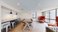 This well-appointed study room offers a thoughtfully designed, single-floor layout that balances open collaboration with private meeting spaces. The main area is anchored by a large conference-style table with ergonomic rolling chairs, set against light hardwood flooring and a textured white ceiling with recessed lighting. Floor-to-ceiling windows flood the space with natural light and provide views of the surrounding courtyard and building exterior, creating an open, airy atmosphere. A tufted leather sofa and bold red high-back lounge chairs offer comfortable informal seating areas for relaxed study or conversation. Along one wall, a sleek kitchenette features an extended granite countertop with built-in power outlets, gray cabinetry, and bar-height seating beneath a statement pendant light — ideal for working meals or casual breaks. Two dedicated private meeting rooms branch off from the main area, each equipped with a wall-mounted television, a whiteboard, and its own conference table and seating. One room includes a round wooden table with bright orange chairs, while the other features a rustic wood table with a window and additional whiteboard space. Together, these elements create a versatile, modern environment well-suited for focused study, group collaboration, and small meetings.