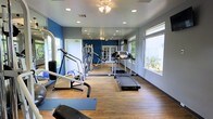 This fitness center is designed to provide a functional and inviting workout environment. The space features warm hardwood flooring throughout, complemented by crisp white walls and bold blue accent walls that give the room a modern, energetic feel. Recessed lighting and a ceiling fan ensure the space remains well-lit and comfortable during workouts. Large windows along the perimeter flood the room with natural light and offer views of the surrounding outdoor area, enhancing the overall atmosphere. A floor-to-ceiling mirror runs along one wall, visually expanding the space and allowing users to monitor their form during exercise. The fitness center is equipped with a variety of cardio machines, including treadmills and an elliptical, as well as strength and resistance training equipment. Exercise mats are available for stretching and floor work. French doors are visible in the mirrored reflection, suggesting a connected space or entryway. The layout is thoughtfully organized to allow for comfortable movement between equipment. Overall, this fitness center offers a clean, well-maintained space that caters to a range of fitness needs in a bright and motivating setting.