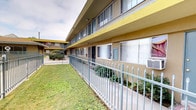 This ground-floor unit is situated within a two-story residential complex characterized by a tan stucco exterior with distinctive yellow accent trim. The building is arranged around a central courtyard, creating a community-oriented layout that promotes accessibility and shared outdoor living.

The unit's exterior features sliding windows with blinds, providing natural light while maintaining privacy. A concrete walkway runs directly along the building's facade, lined with low-maintenance landscaping including ornamental grasses and flowering plants, adding greenery along the path to each entry door. The teal-colored unit doors provide a visual contrast against the neutral stucco walls.

The complex includes shared amenities, most notably a fenced swimming pool visible from the courtyard area, surrounded by mature shrubs and trees. A metal staircase with aggregate-textured steps connects the ground level to the upper floor, featuring a sturdy metal railing. An iron perimeter fence borders the grassy common area, providing both security and a defined boundary.

Overall, the complex offers a functional, well-maintained environment with accessible outdoor spaces and practical amenities suited for everyday residential living.