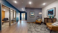 This thoughtfully designed single-floor office space combines contemporary aesthetics with functional layouts to create an engaging and professional environment. The reception and lounge area immediately sets the tone with bold teal accent walls, textured gray wallcovering, and warm wood-look flooring that transitions seamlessly into patterned carpet. A pair of mid-century modern mustard-yellow armchairs, a geometric area rug, and curated framed artwork lend the space a sophisticated yet welcoming character. A wooden credenza with decorative accents adds a refined finishing touch to the lounge. The corridor connects to a dedicated conference room featuring a striking burnt-orange accent wall, large windows that invite abundant natural light and exterior views, a drop ceiling with recessed and panel lighting, and a full-sized conference table surrounded by ergonomic mesh chairs—ideal for productive team meetings. Adjacent to the conference room is a compact, private meeting nook, also finished with a vibrant orange accent wall, comfortable modern lounge chairs, side tables, and bold abstract artwork, providing an intimate setting for focused conversations. Throughout the space, consistent use of colorful accents, curated furnishings, and varied flooring materials create a cohesive, energetic atmosphere suited to a modern professional setting.