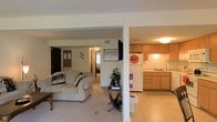 This single-floor residence is designed with an open-concept layout that integrates the living and kitchen areas into one cohesive space. A structural column subtly defines the boundary between the two zones while maintaining a sense of openness throughout. The living area features plush, neutral-toned carpeting and is furnished with a comfortable sofa accented with decorative pillows, centered around a wall-mounted flat-screen television. A small side table with a potted plant adds a touch of greenery to the space. The kitchen is positioned adjacent to the living area and is equipped with light wood cabinetry, a white refrigerator, and a single-basin sink beneath overhead task lighting. The cabinetry provides ample storage, and the layout supports efficient use of the cooking space. A hallway visible from the main living area leads further into the residence, hinting at additional rooms beyond, with a glimpse of natural light filtering through from a window at the far end. The overall ambiance is warm and practical, with consistent neutral tones in the walls, flooring, and furnishings creating a unified and inviting atmosphere throughout the home.